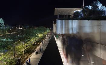 Photo taken at night which shows people walking along a path that is next to trees and a lake and behind them is a large, glass building